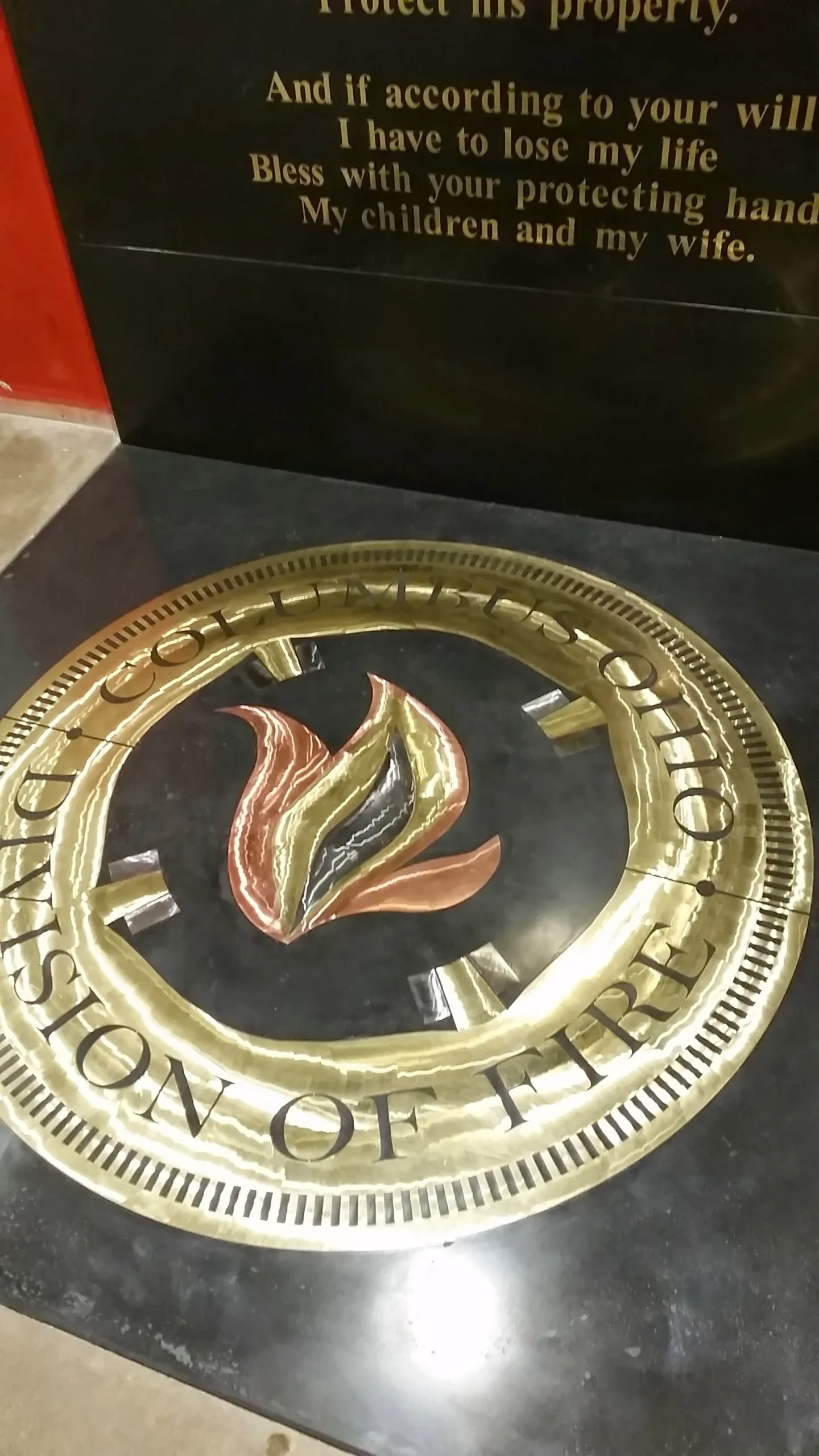 A brass circular plaque with engraved text around the rim displaying a stylized flame design in red, gold, and black colors in the center, mounted on a dark display board with inspirational text above it