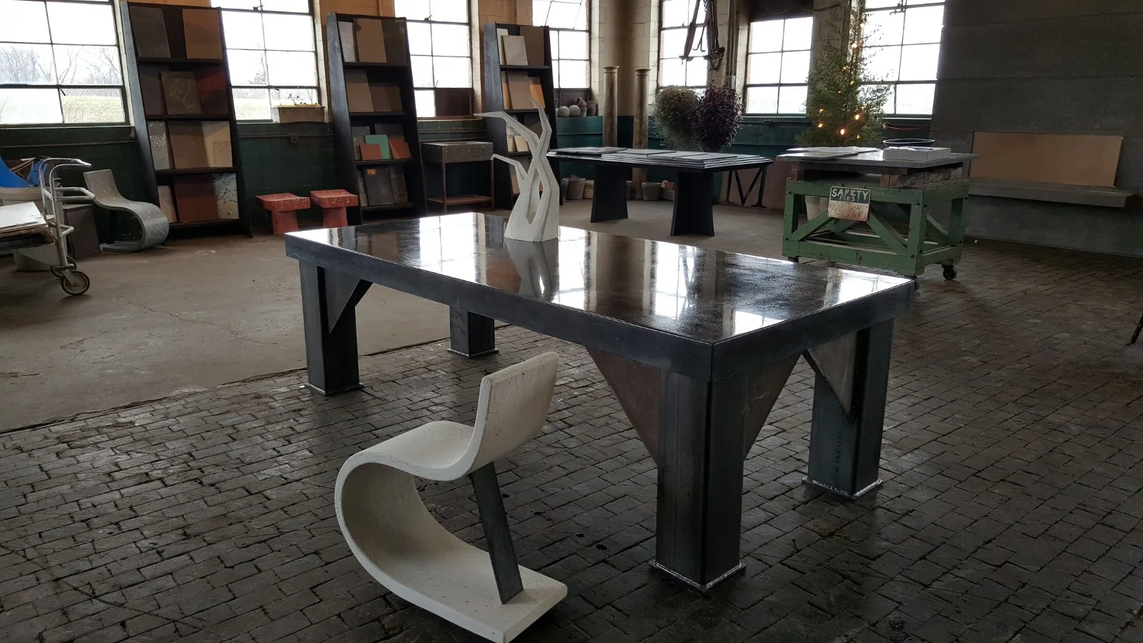 Modern furniture showroom with sleek black dining table, white sculptural chairs, shelving units, and industrial windows in contemporary warehouse space