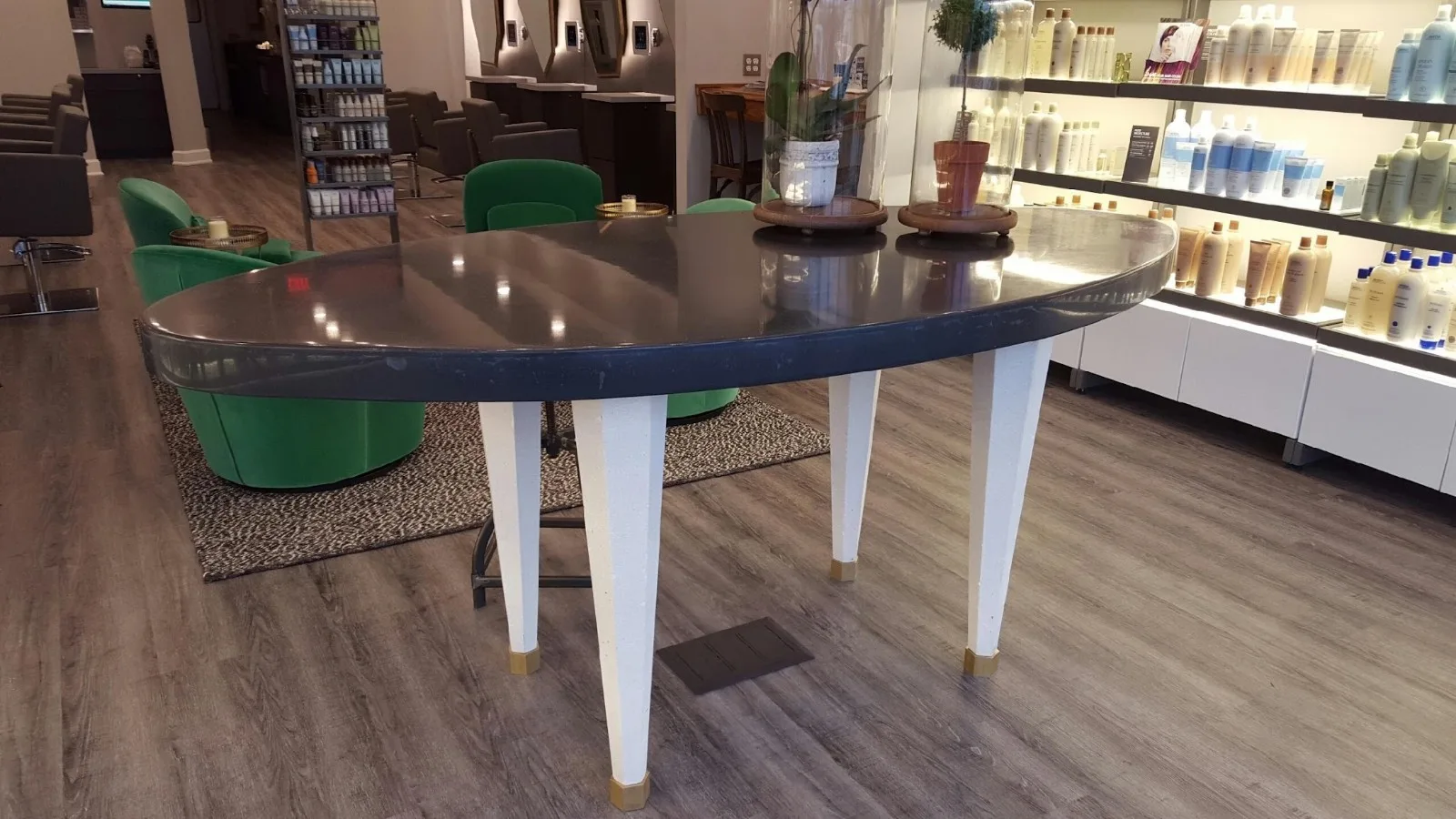 Modern salon reception area with curved dark table, white support legs, green chairs, and illuminated product shelves displaying hair care bottles
