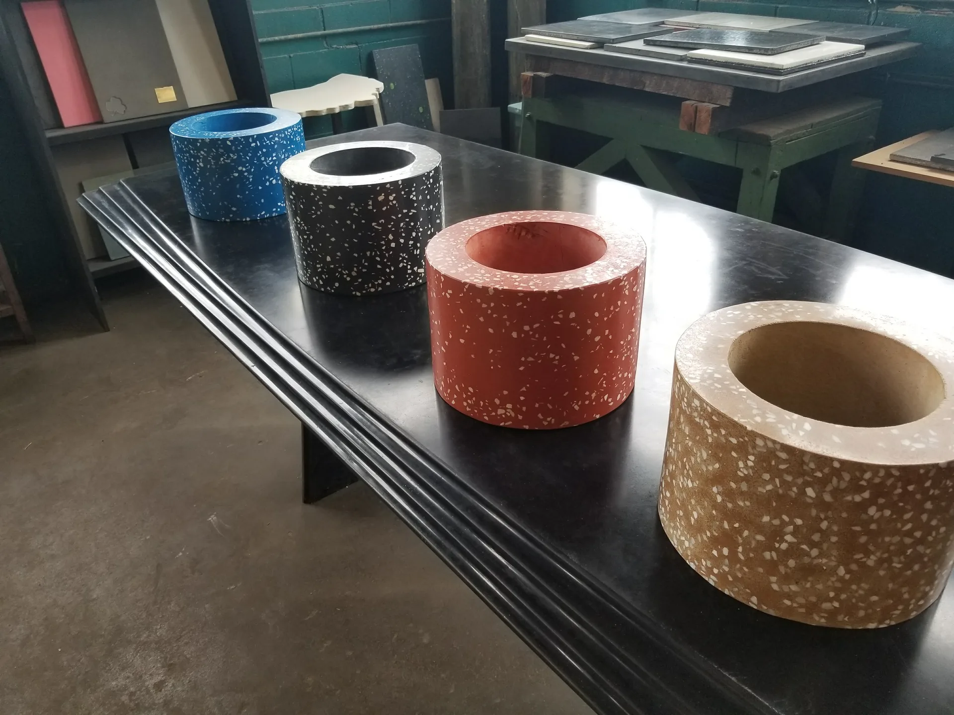 Five cylindrical concrete planters with terrazzo aggregate in blue, black, terracotta, and tan displayed on a metal table in an industrial workspace
