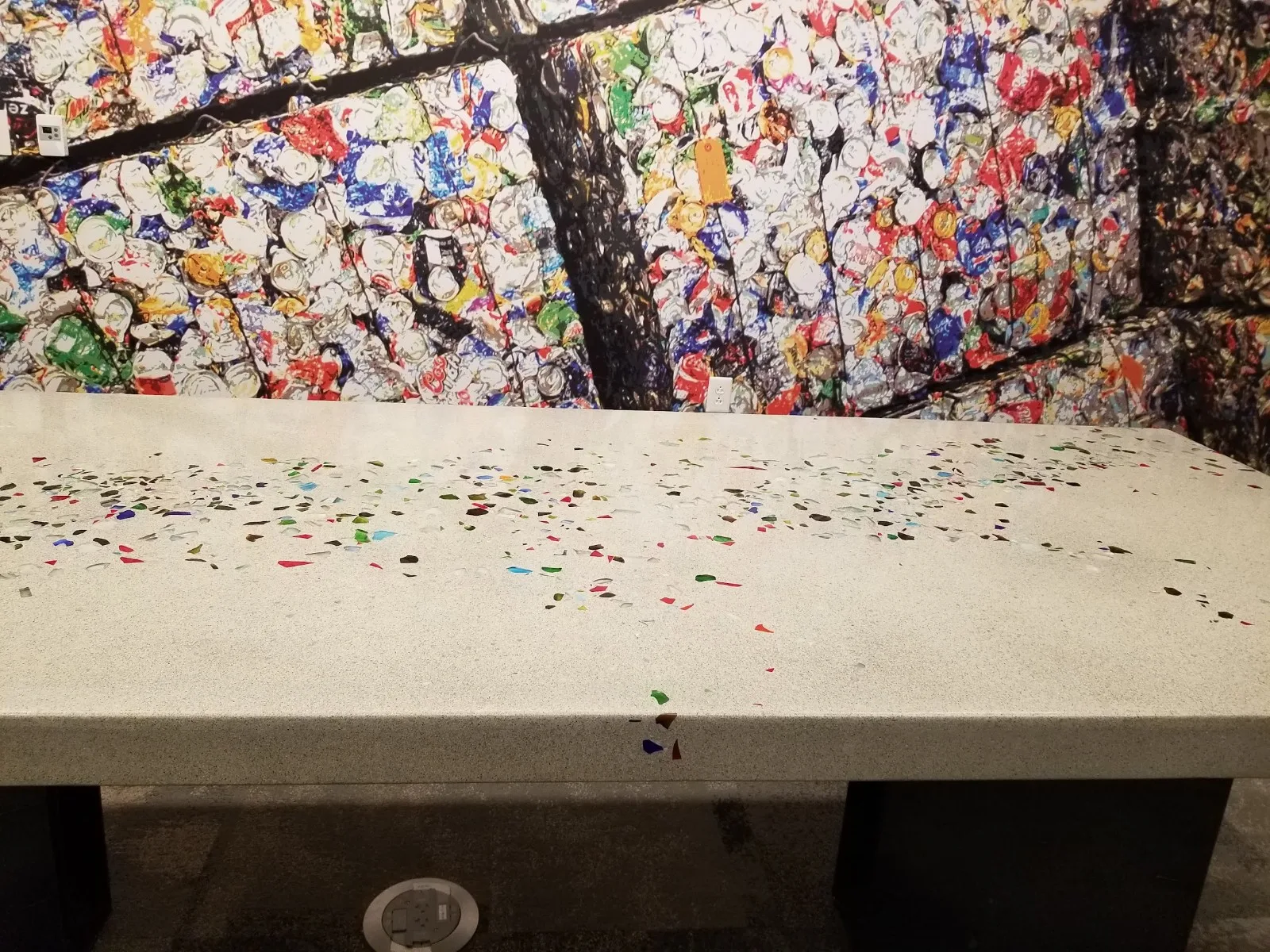 Compressed bales of crushed aluminum cans stacked against warehouse wall with colorful debris scattered on concrete floor below