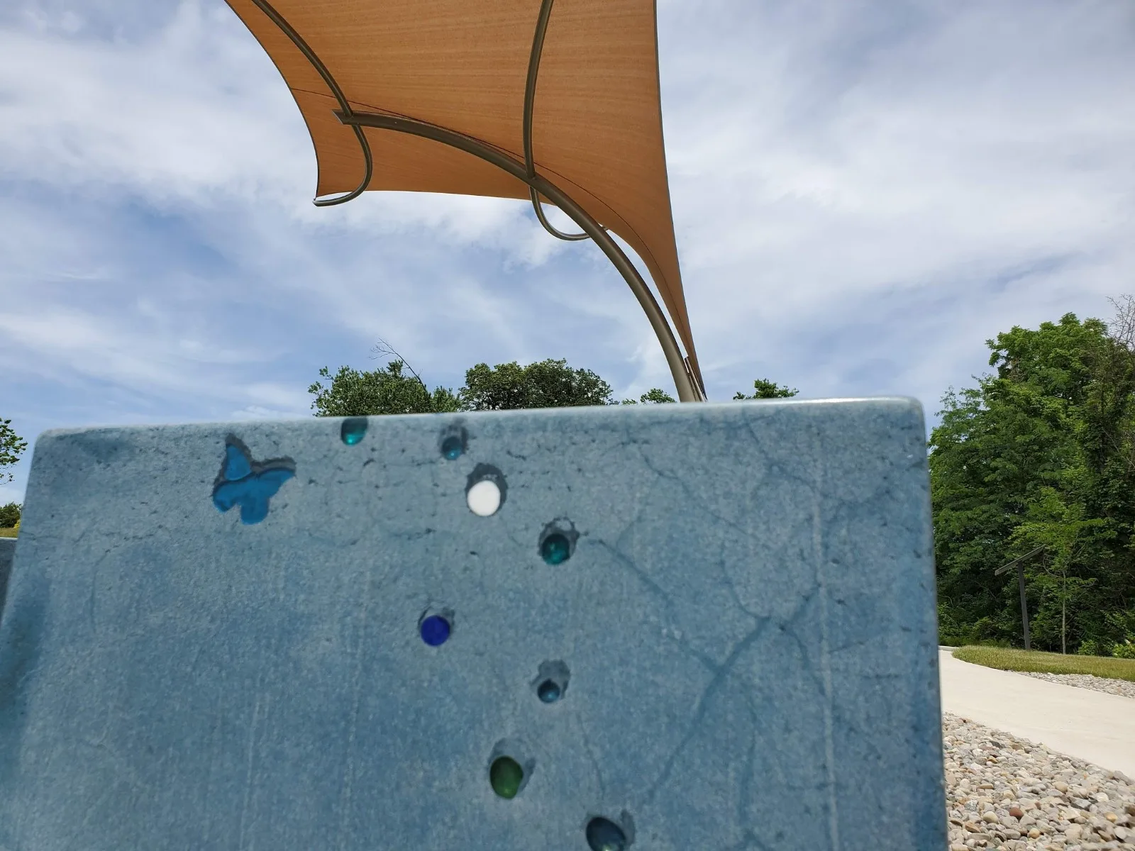 Blue playground climbing wall with colorful holds and a butterfly design under a tan shade sail, with trees visible in background
