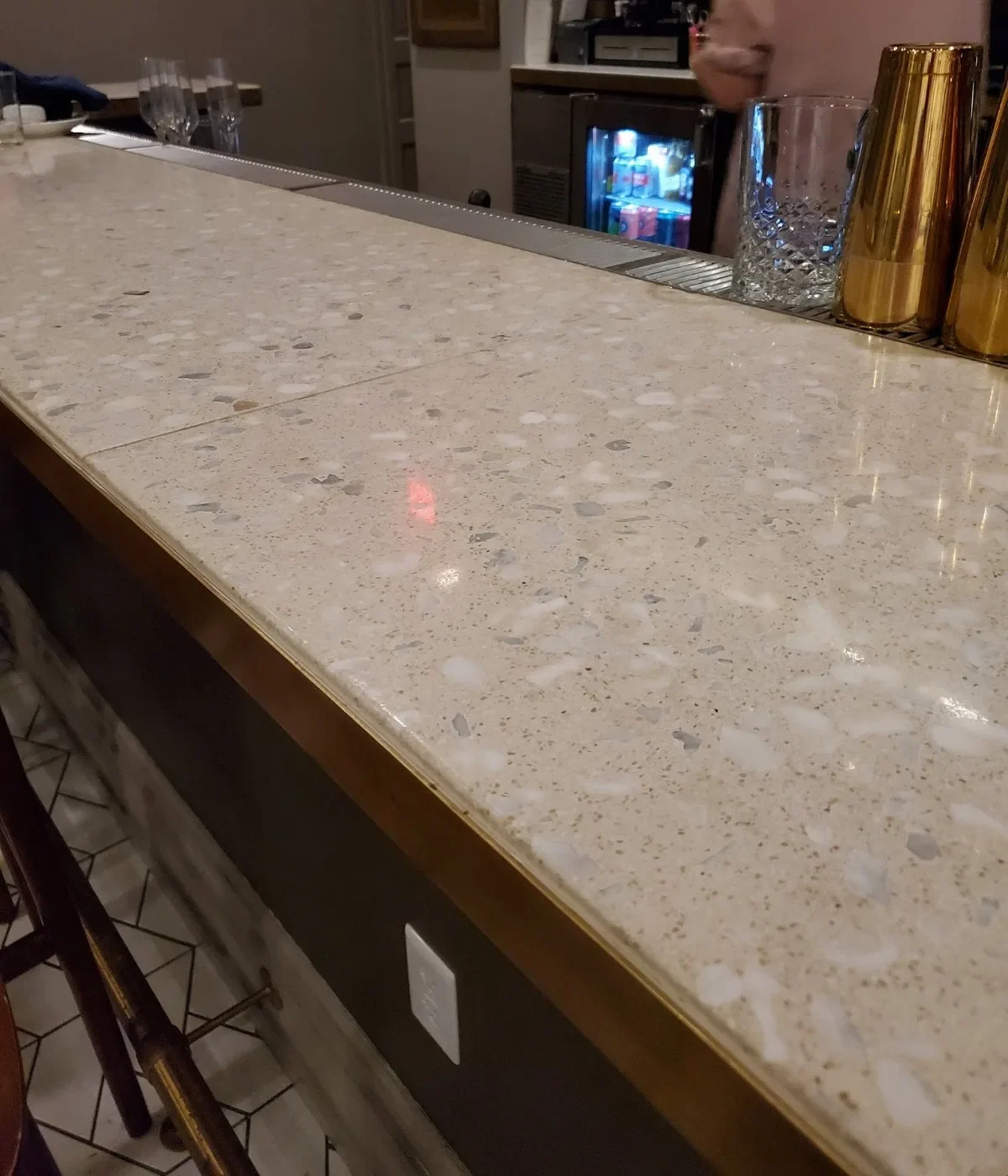Marble countertop at a bar or reception area with glassware and gold container, person in pink shirt standing behind counter with computer screens visible in background