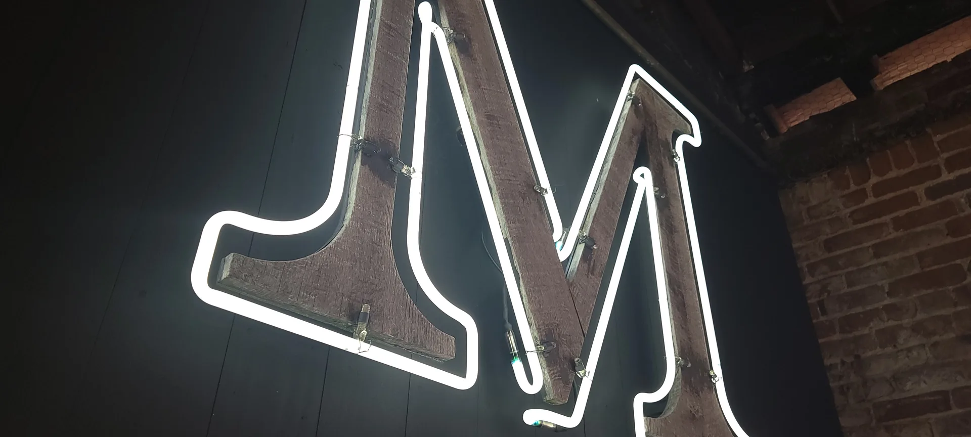 Glowing white neon sign with stylized letters mounted on wooden beams against dark background
