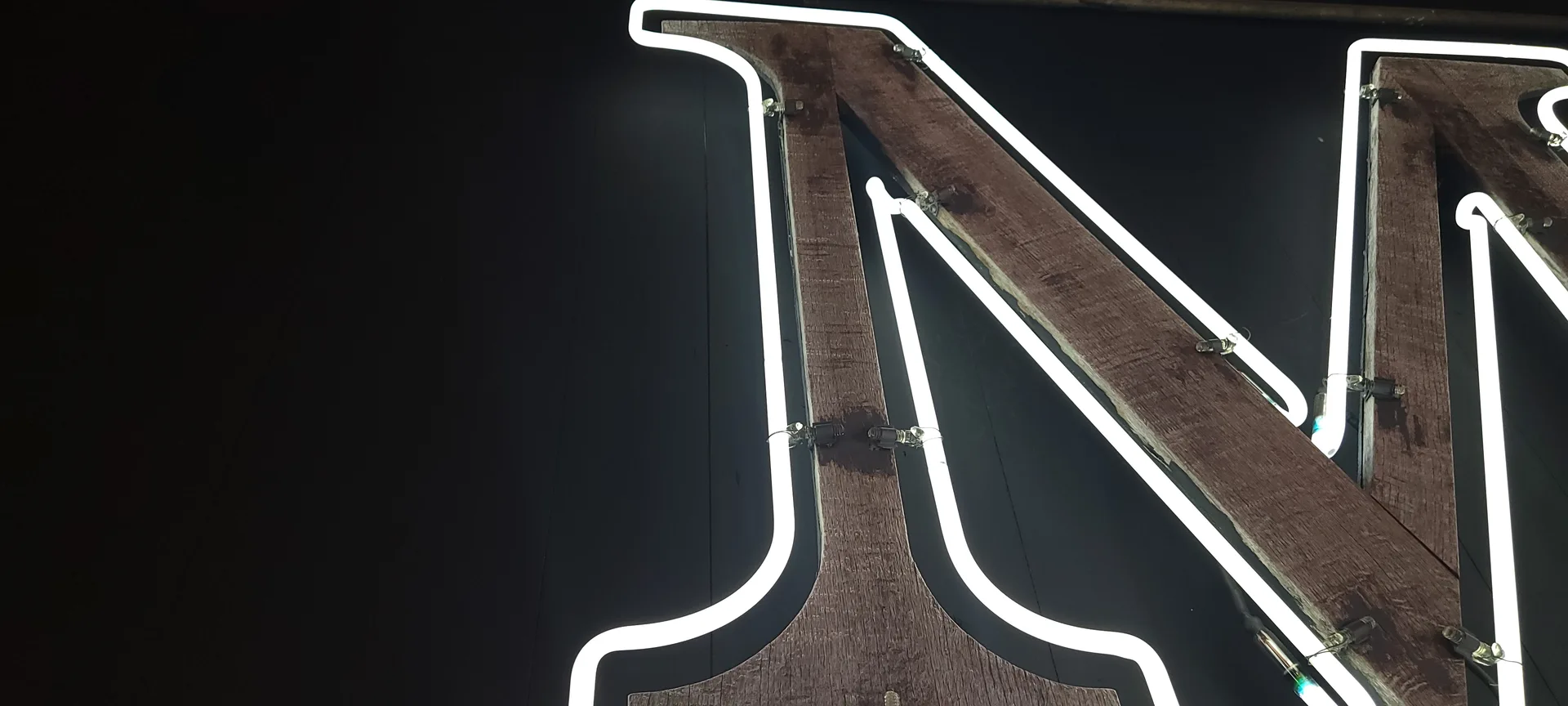 Neon sign with white glowing tubes and dark wooden frame against black background, featuring angular geometric design with curved and straight lines