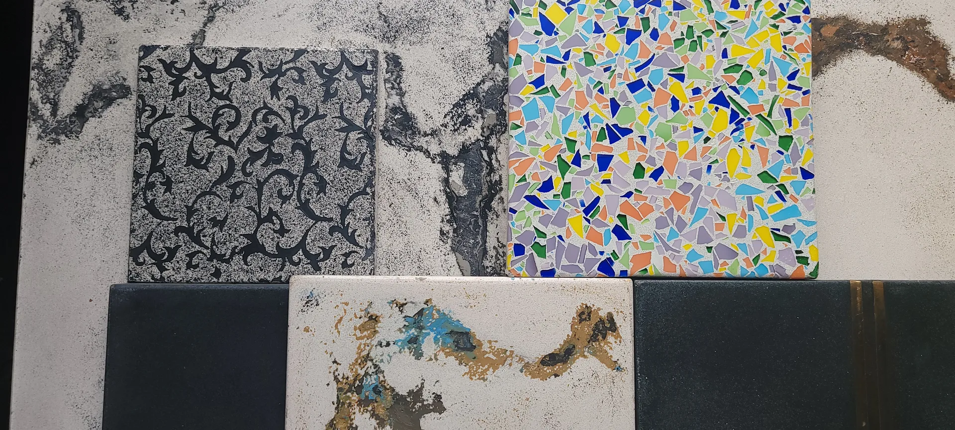 Five material samples arranged on concrete surface: ornate black pattern on textured gray, vibrant mosaic tile pieces in multiple colors, plain black swatches, and weathered cream tile with blue and gold distressing
