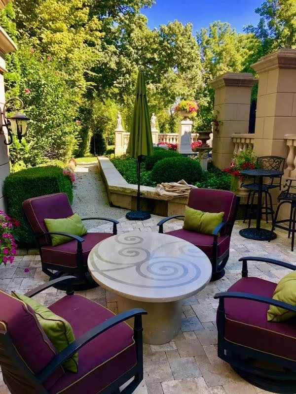 Luxurious backyard patio with purple cushioned chairs arranged around a decorative circular stone table, surrounded by manicured hedges, tall trees, and flowering plants under clear blue sky