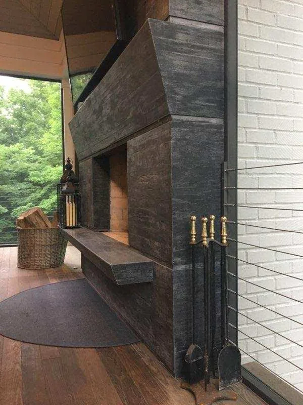Modern dark slate fireplace with metal andirons and brass accents in contemporary living space with large window overlooking trees