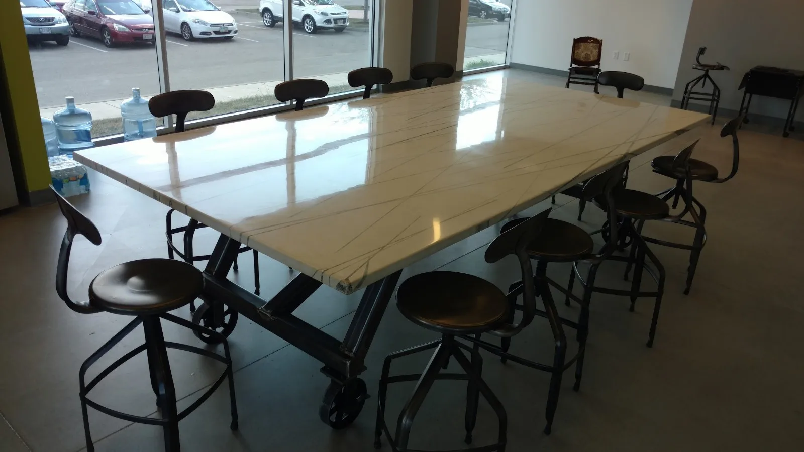 Modern conference room with long light wood table, black chairs with wheels, large windows overlooking parking lot with cars, water dispensers, and additional seating in background