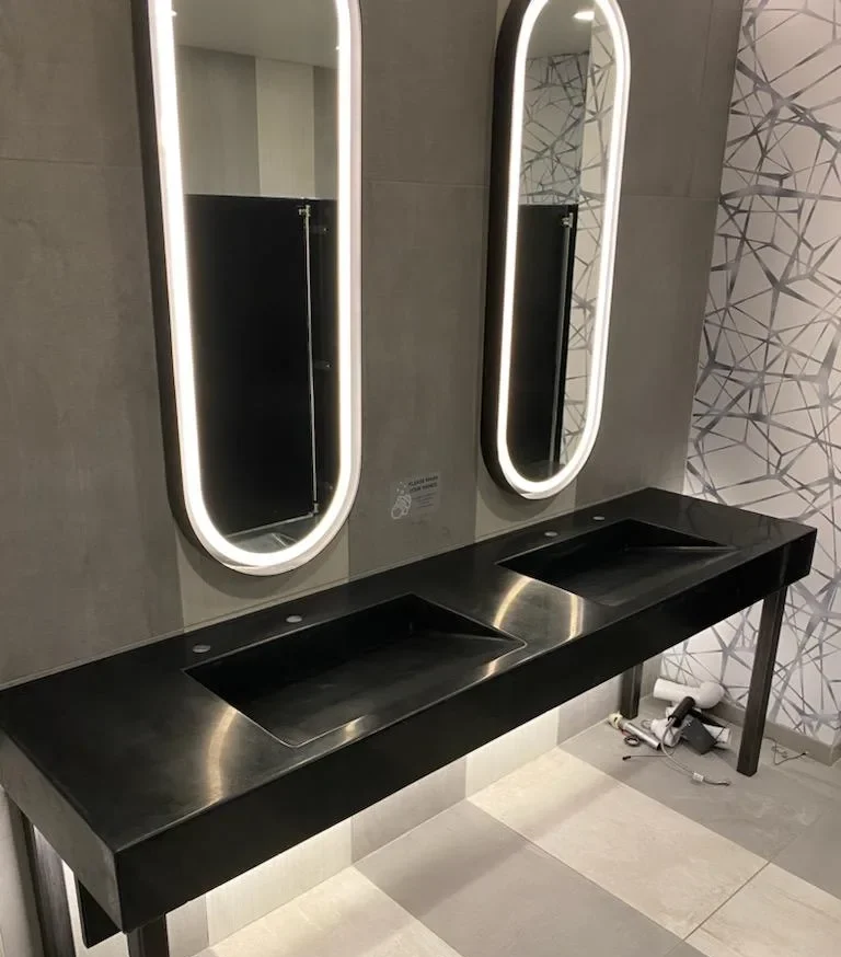 Modern commercial bathroom with black countertop sink and two illuminated oval mirrors above, geometric patterned wall tile visible on right side
