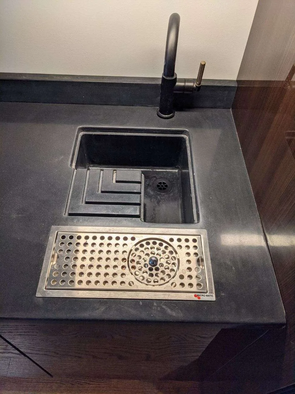 Black commercial bar sink with faucet, drain grate, and ice bin compartment built into dark countertop