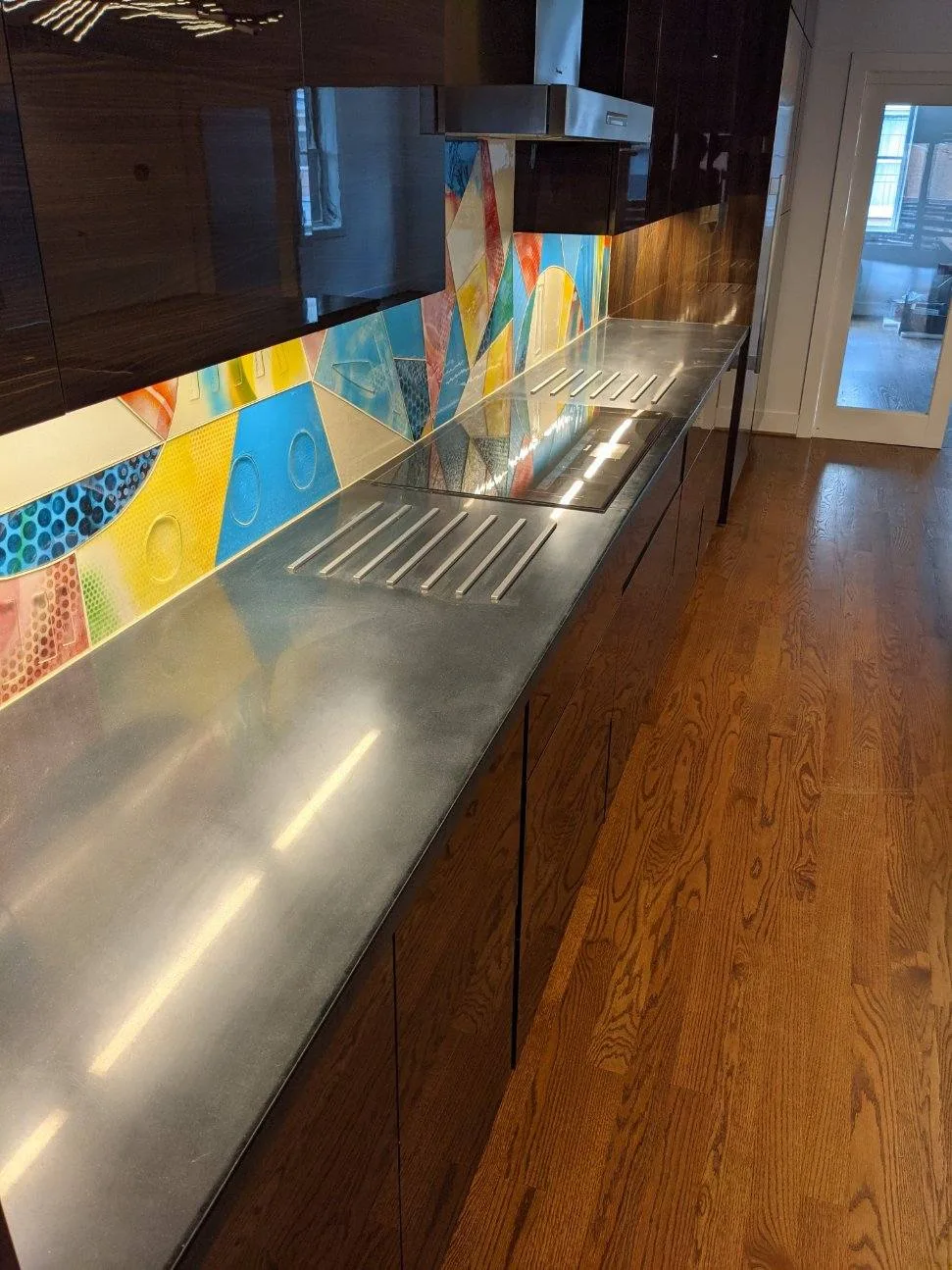 Modern kitchen with stainless steel countertops, colorful geometric backsplash tile in blue, yellow, and orange, dark wood cabinetry, and hardwood flooring