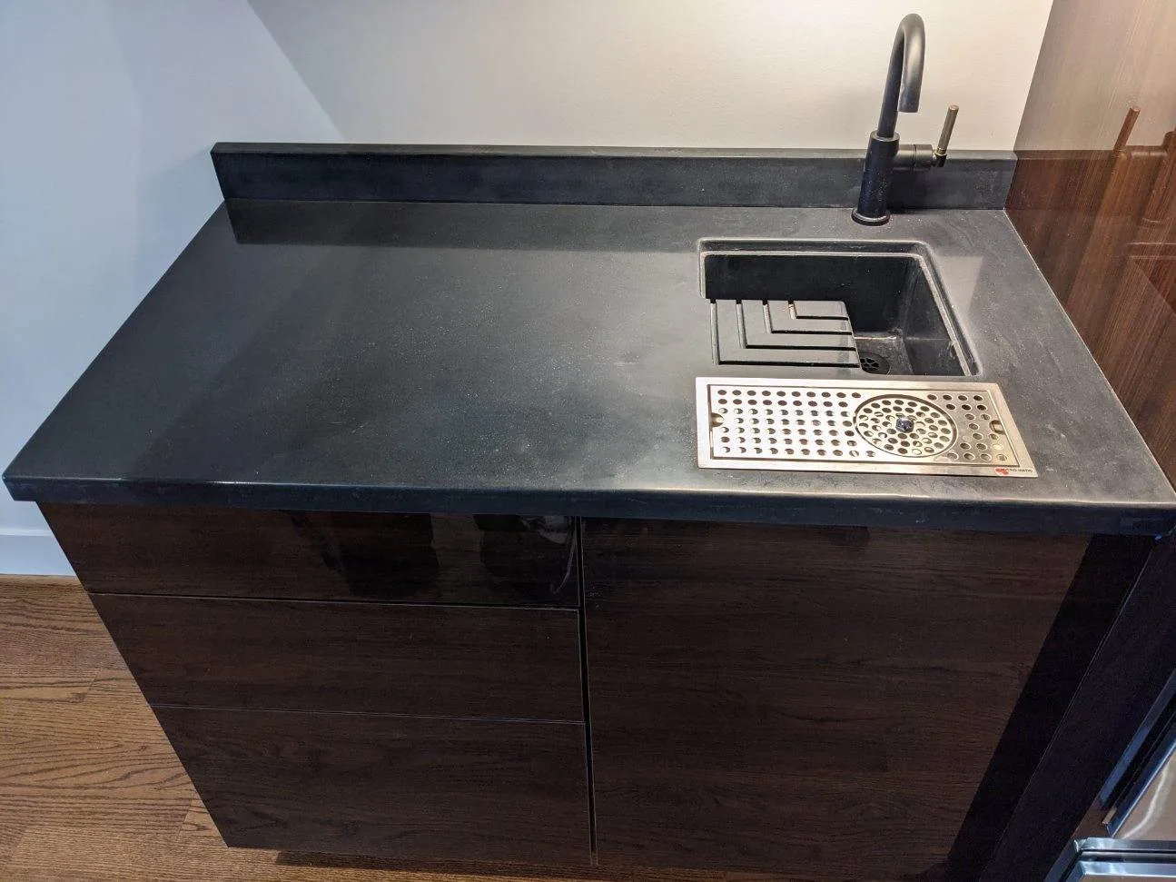 Dark wood bar sink unit with black countertop, stainless steel drainboard, and black faucet against white wall