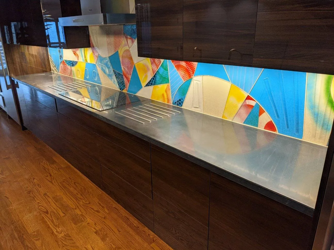 Modern kitchen countertop with stainless steel surface and colorful geometric backsplash featuring blue, yellow, red and green triangular patterns with dark cabinetry above