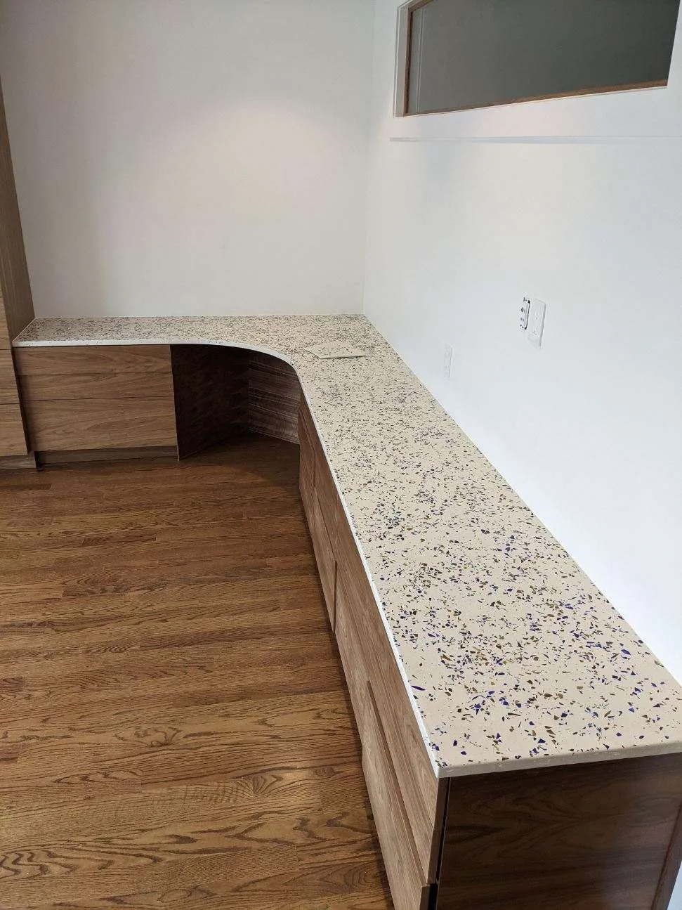 Empty office space with L-shaped desk featuring speckled countertop, wood cabinetry, hardwood flooring, and white walls with window