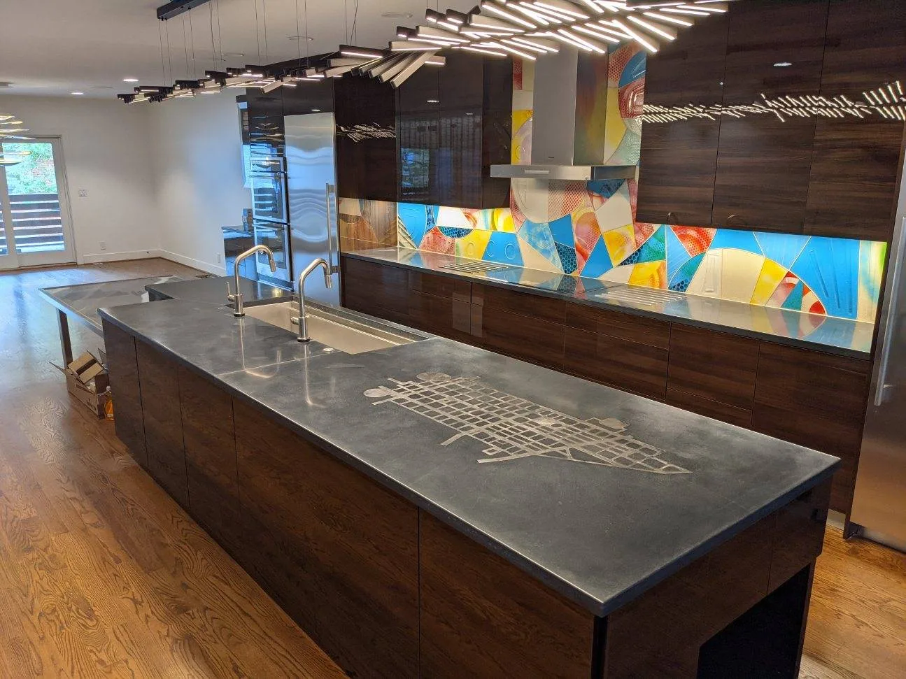 Modern kitchen interior with dark wood cabinetry, gray concrete countertops, colorful geometric backsplash, stainless steel sink fixtures, and linear pendant lighting overhead