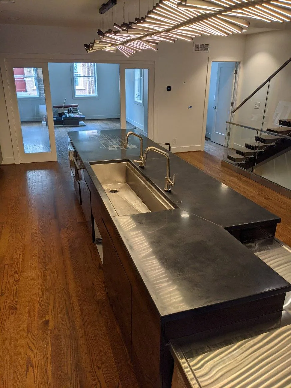 Modern kitchen with stainless steel countertops and sink, dark wood cabinetry, hardwood flooring, and contemporary linear ceiling lights in an open-concept loft space with glass partitions