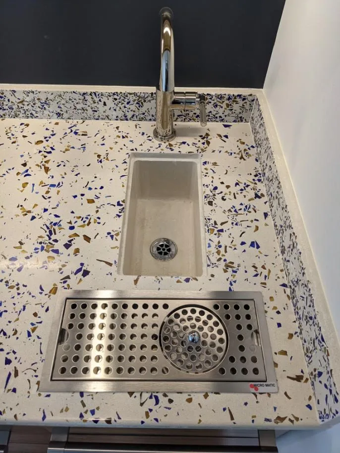 Modern bathroom sink with terrazzo countertop featuring blue and gold flecks, chrome faucet, and stainless steel drainboard with circular drain detail