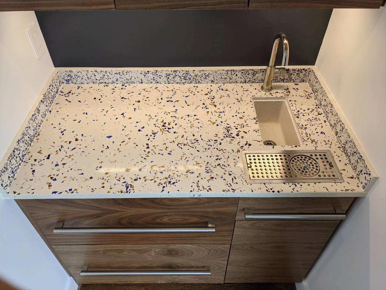 Modern kitchenette with terrazzo countertop, stainless steel sink, faucet, and drain board next to dark wood cabinetry with metal handles