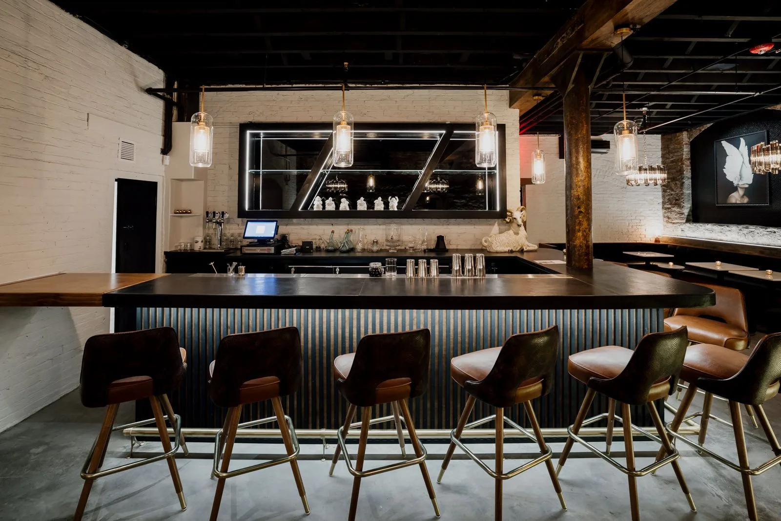 Modern bar interior with black counter, wooden mid-century chairs, pendant lights, and illuminated shelving displaying bottles against white brick walls