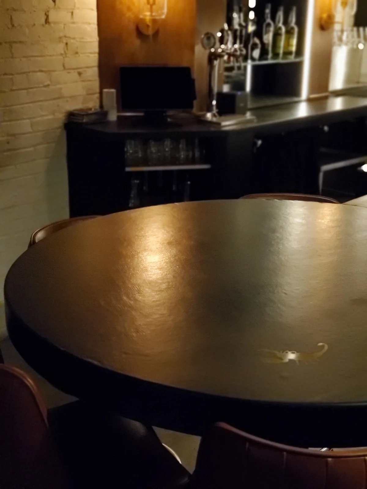 Round metallic table in foreground with bar counter and shelving displaying bottles in background of dimly lit establishment