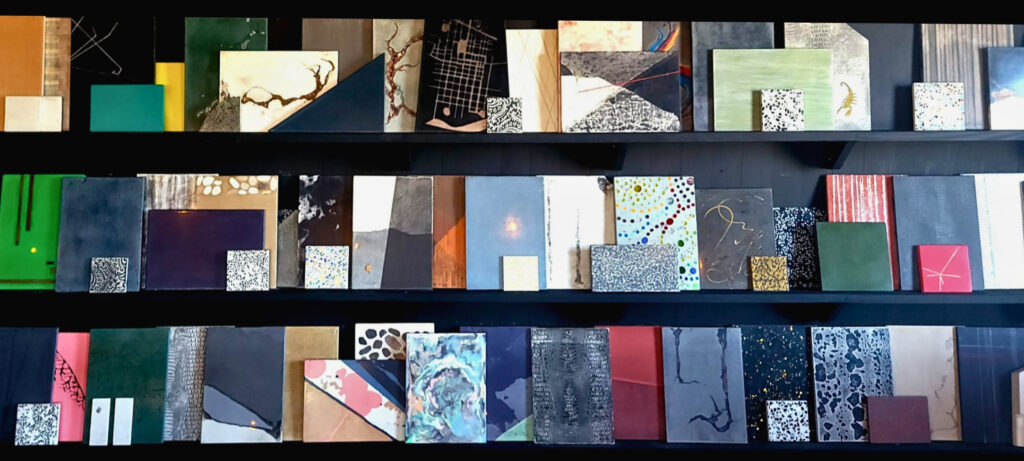 Three shelves displaying an organized collection of design material samples, color swatches, and textured papers in navy, teal, pink, gold, and neutral tones with various patterns and finishes
