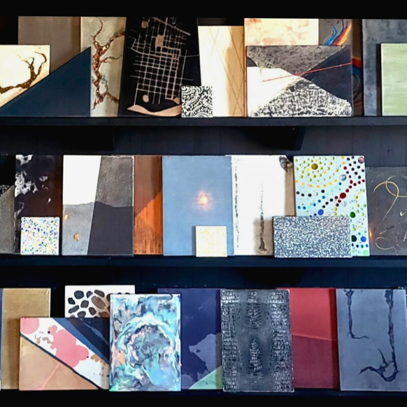 Three shelves displaying an organized collection of design material samples, color swatches, and textured papers in navy, teal, pink, gold, and neutral tones with various patterns and finishes