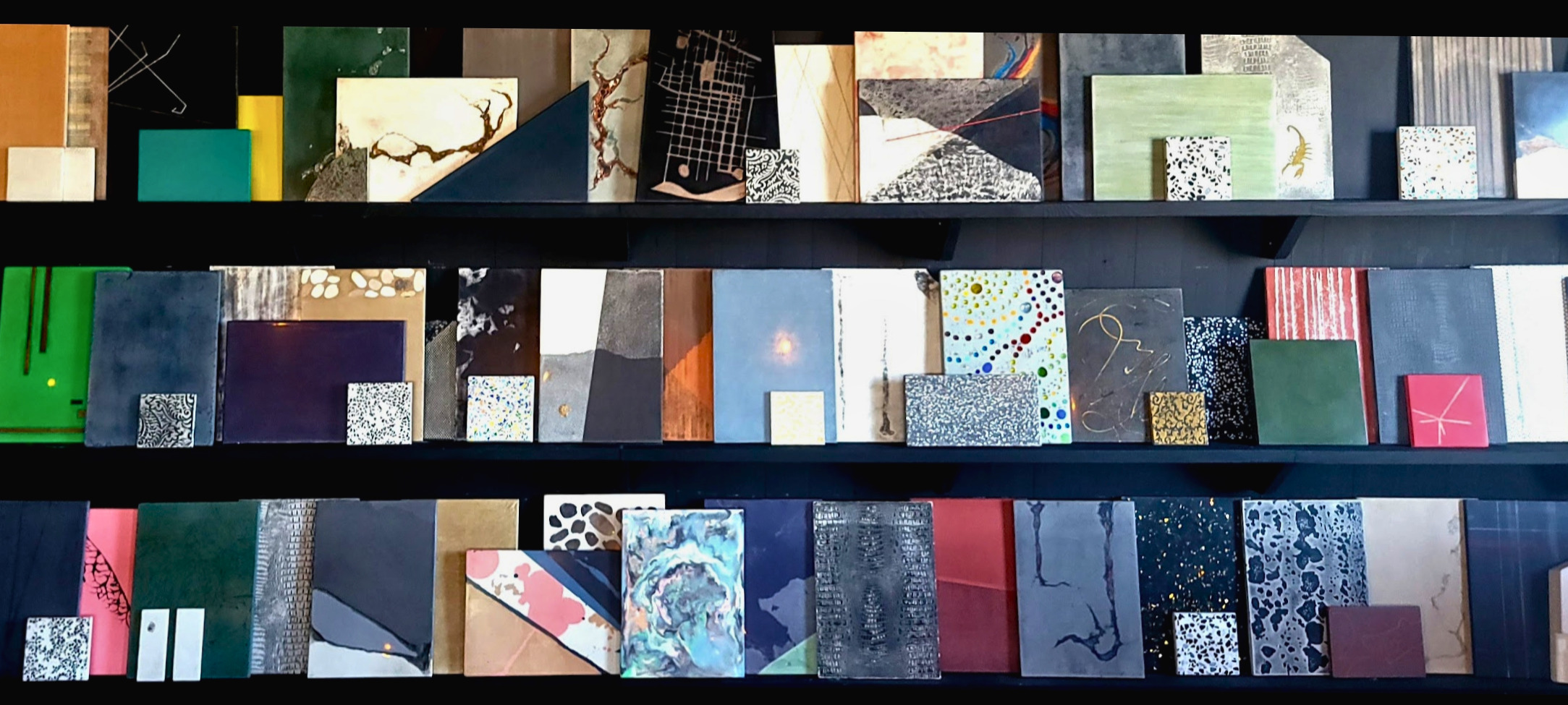 Three shelves displaying an organized collection of design material samples, color swatches, and textured papers in navy, teal, pink, gold, and neutral tones with various patterns and finishes