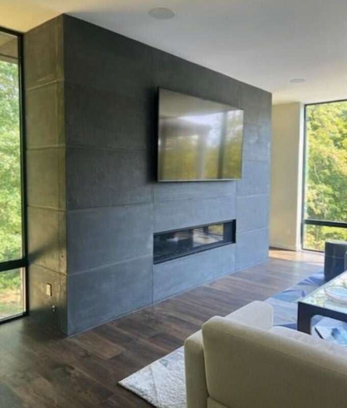Modern living room with dark slate tile fireplace wall, mounted flat-screen TV, linear fireplace insert, dark wood flooring, cream armchair, and floor-to-ceiling windows overlooking green landscape