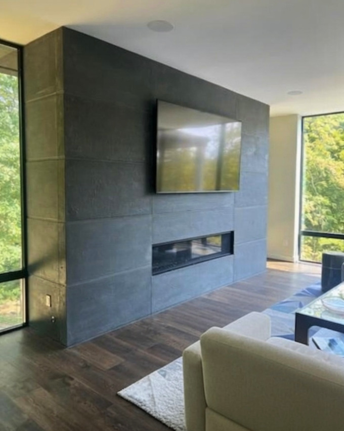 Modern living room with dark slate tile fireplace wall, mounted flat-screen TV, linear fireplace insert, dark wood flooring, cream armchair, and floor-to-ceiling windows overlooking green landscape