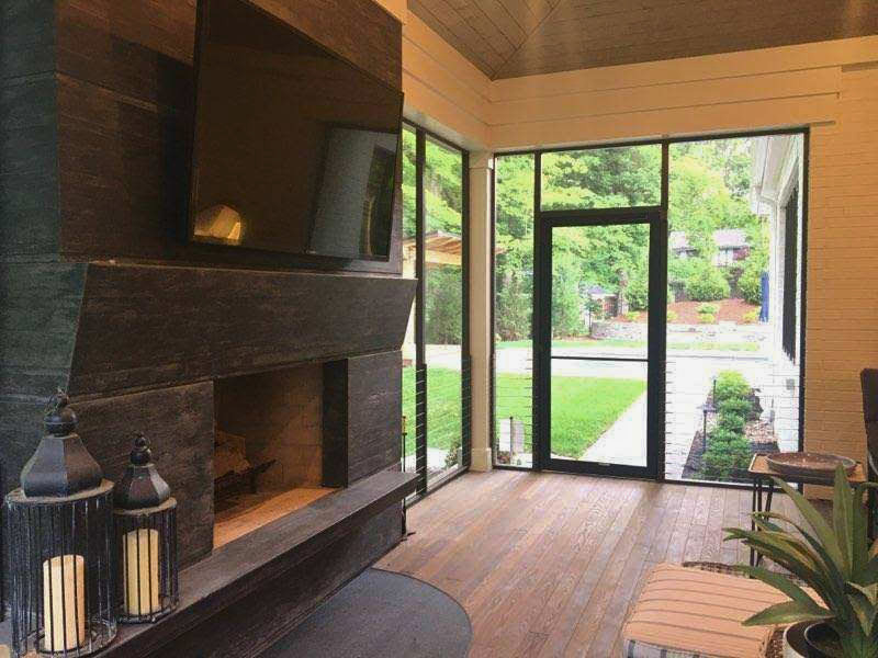 Modern patio room with dark fireplace, wooden floors, and glass doors opening to lush green garden views