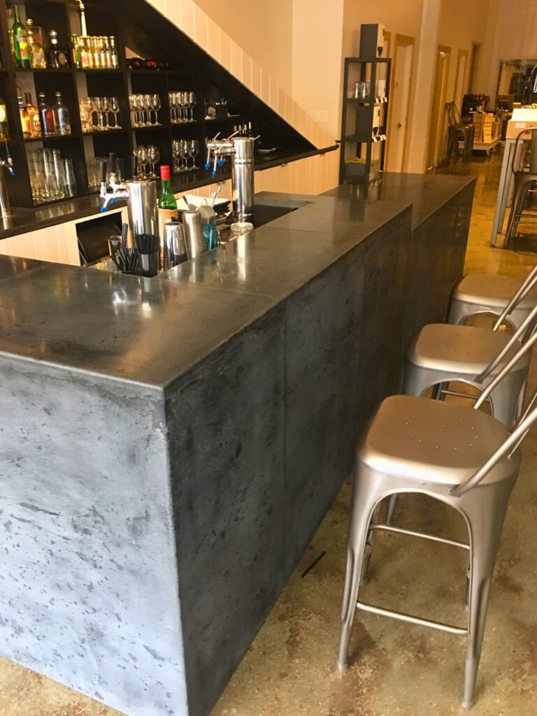 Modern bar counter with concrete finish, metal bar stools, shelving with liquor bottles and glassware, and stainless steel tap handles in an upscale venue