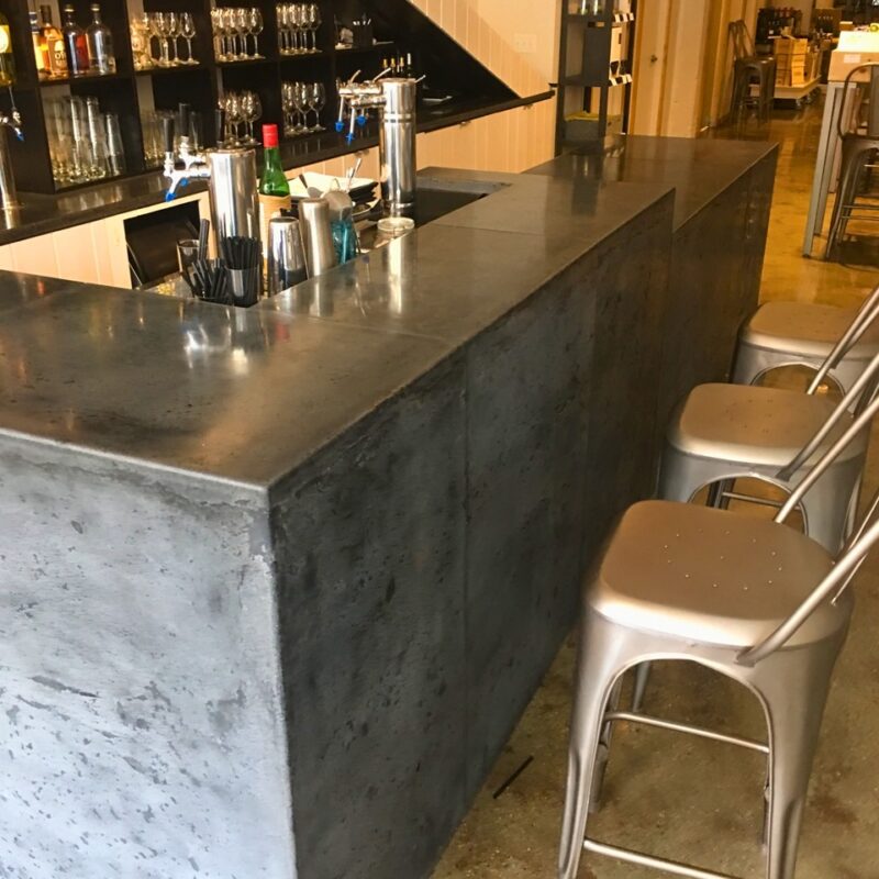 Modern bar counter with concrete finish, metal bar stools, shelving with liquor bottles and glassware, and stainless steel tap handles in an upscale venue