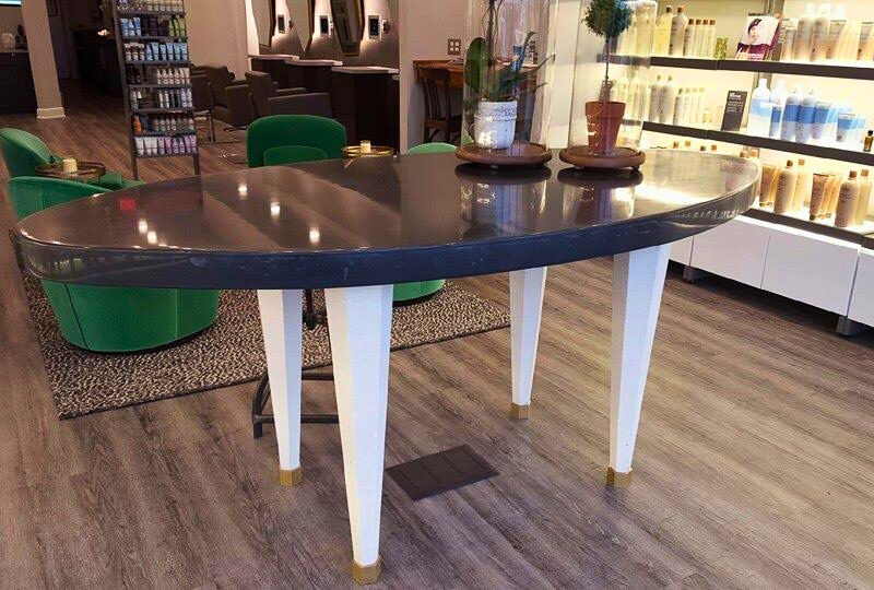 Modern salon interior with curved dark table, green chairs, white legs, wooden flooring, and illuminated product shelves displaying hair care bottles