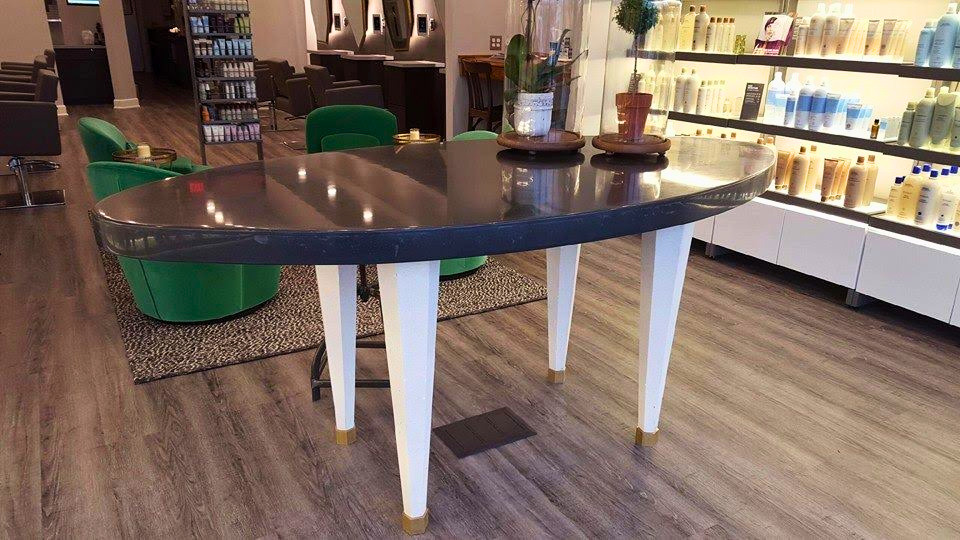 Modern salon interior with curved dark table, green chairs, white legs, wooden flooring, and illuminated product shelves displaying hair care bottles
