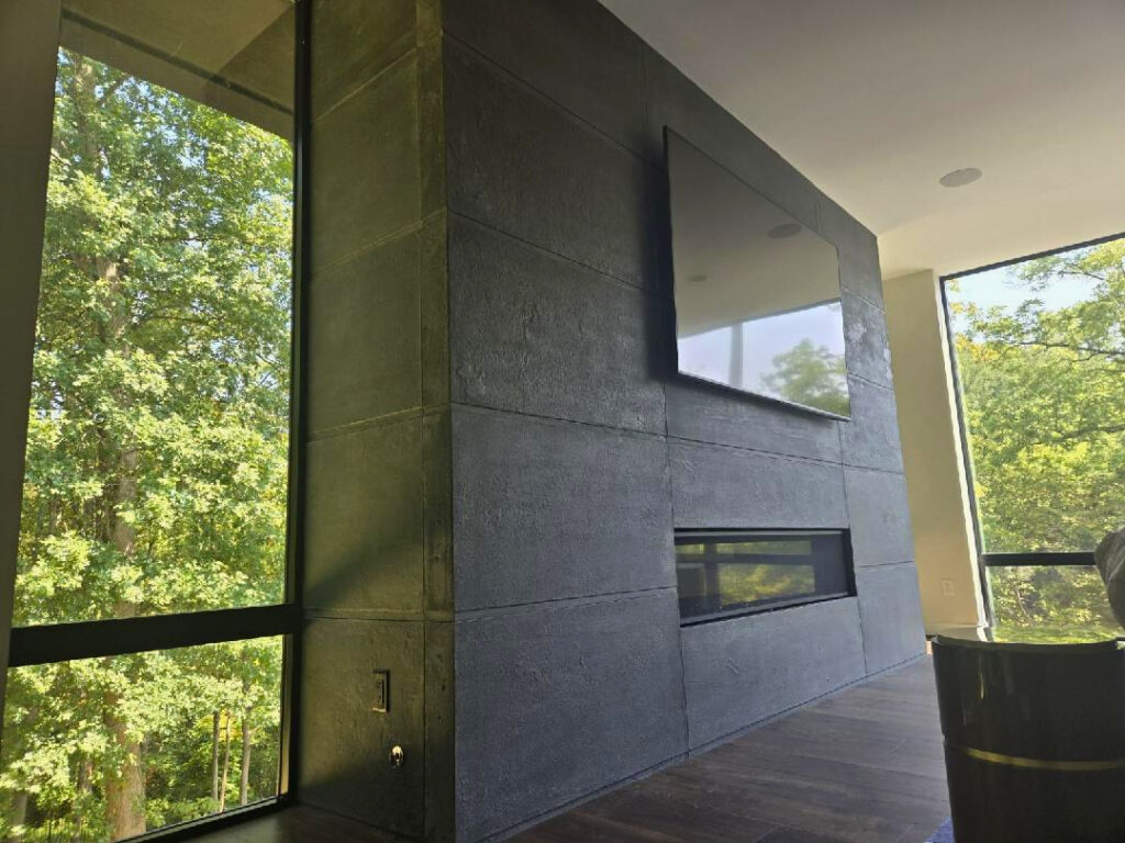 Modern living room with floor-to-ceiling windows overlooking trees, dark textured accent wall with mounted TV and linear fireplace, wooden flooring, and minimalist design