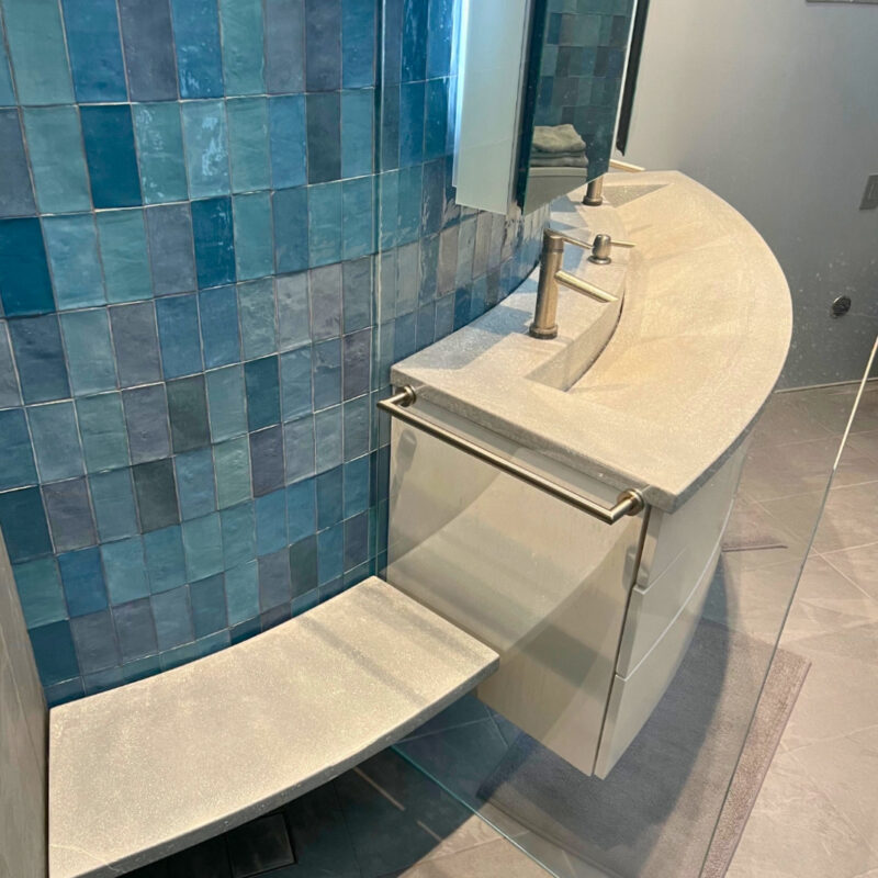 Modern bathroom with curved tan countertop sink, dual faucets, and blue tile shower enclosure with glass panel