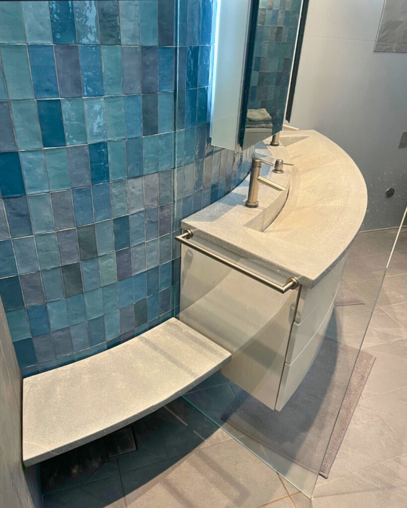 Modern bathroom with curved tan countertop sink, dual faucets, and blue tile shower enclosure with glass panel
