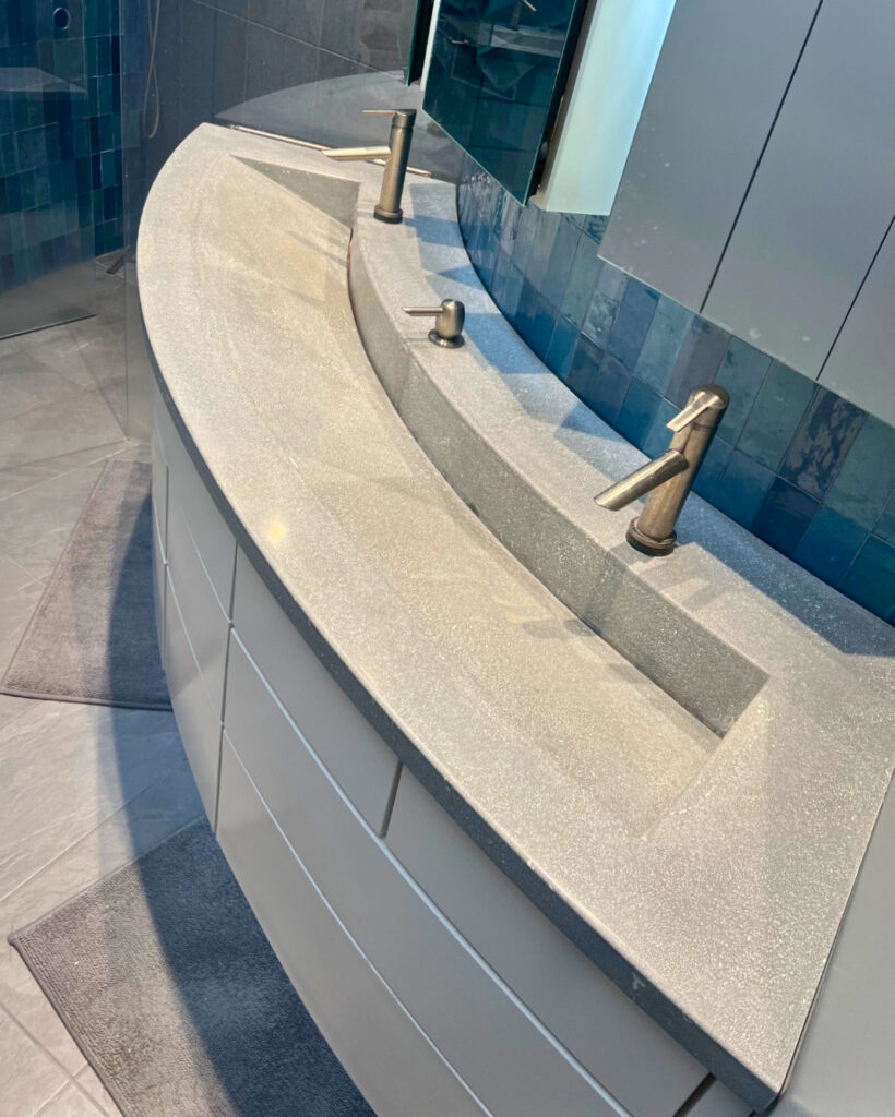 Modern bathroom vanity with curved beige countertop, dual integrated sinks, bronze faucets, and teal tile backsplash