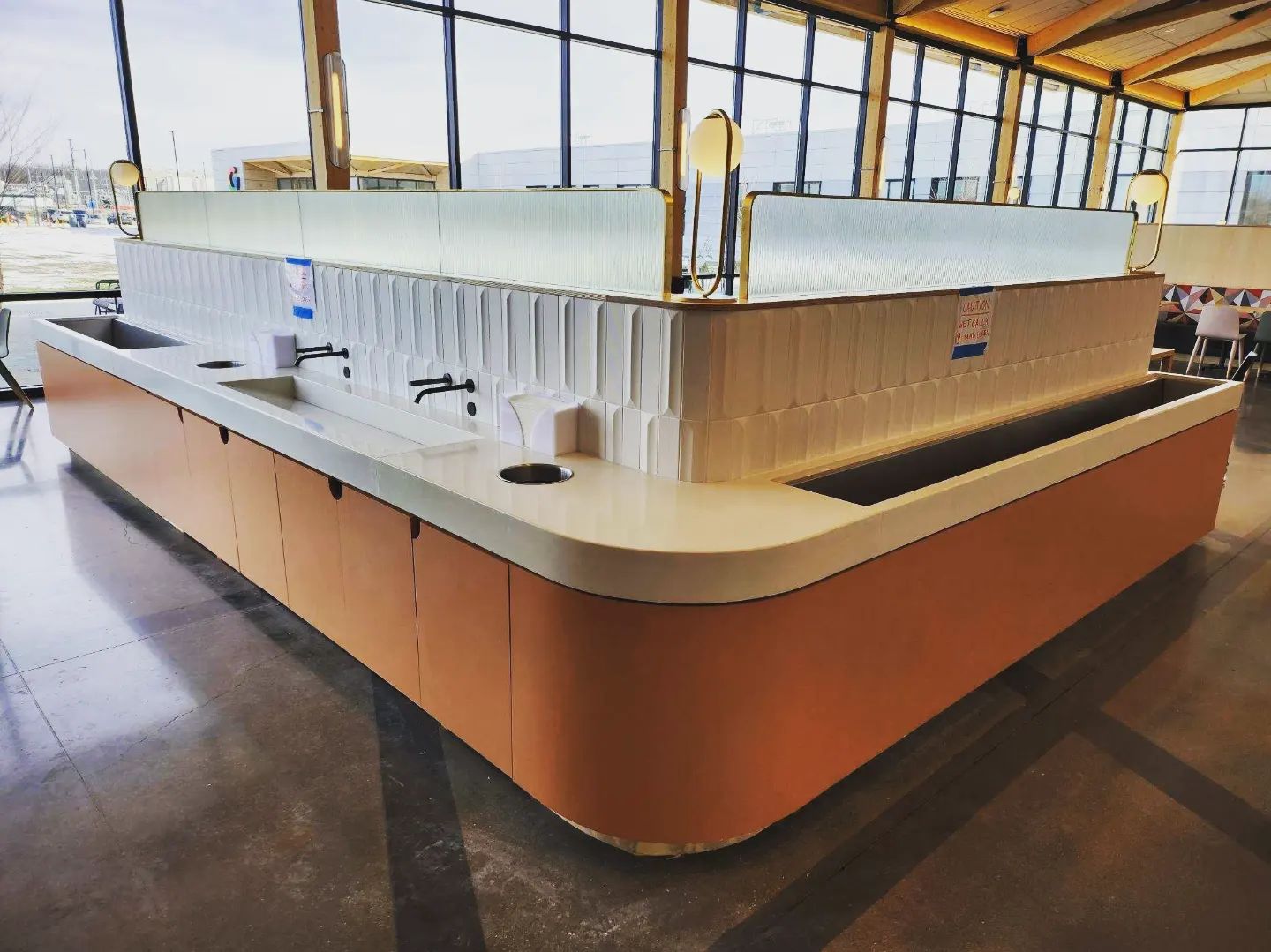 Modern restaurant counter with orange base and cream-colored top, featuring white vertical dividers and black faucets, large windows overlooking marina in background