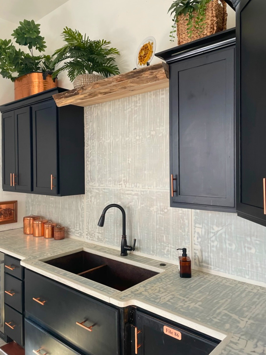 Modern black kitchen with white textured backsplash, copper accents, live edge wooden shelf, potted plants, and black undermount sink