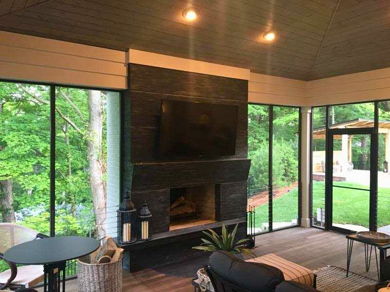 Modern sunroom with black fireplace, comfortable seating, wooden ceiling, and floor-to-ceiling windows overlooking wooded backyard