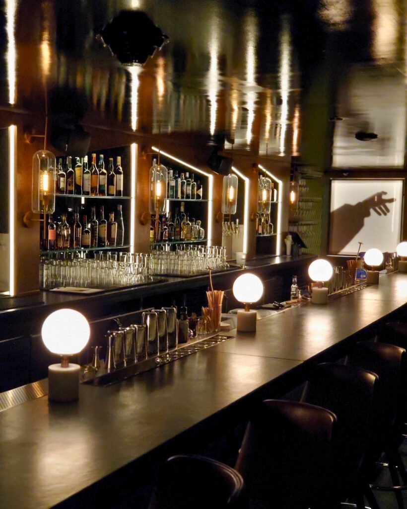 Upscale bar interior with illuminated bottle display, glowing orb lights along counter, brass accents, and modern artwork on walls