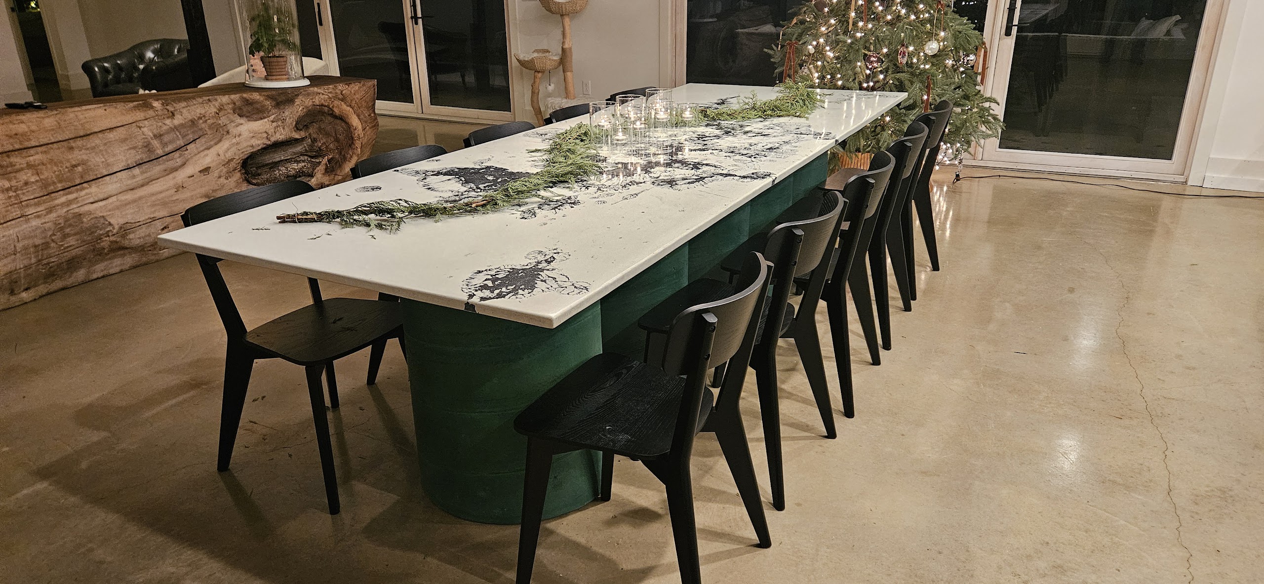 Modern dining table with white and black marble-look top, forest green upholstered base, and black chairs arranged in a bright open room with large windows and potted plants