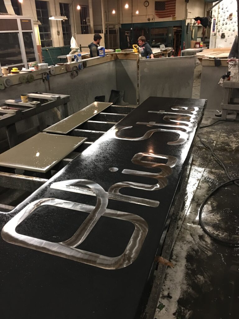 Metal fabrication workshop with stainless steel handles and brackets laid out on a black table, two workers visible at stations in background, Cincinnati industrial workspace