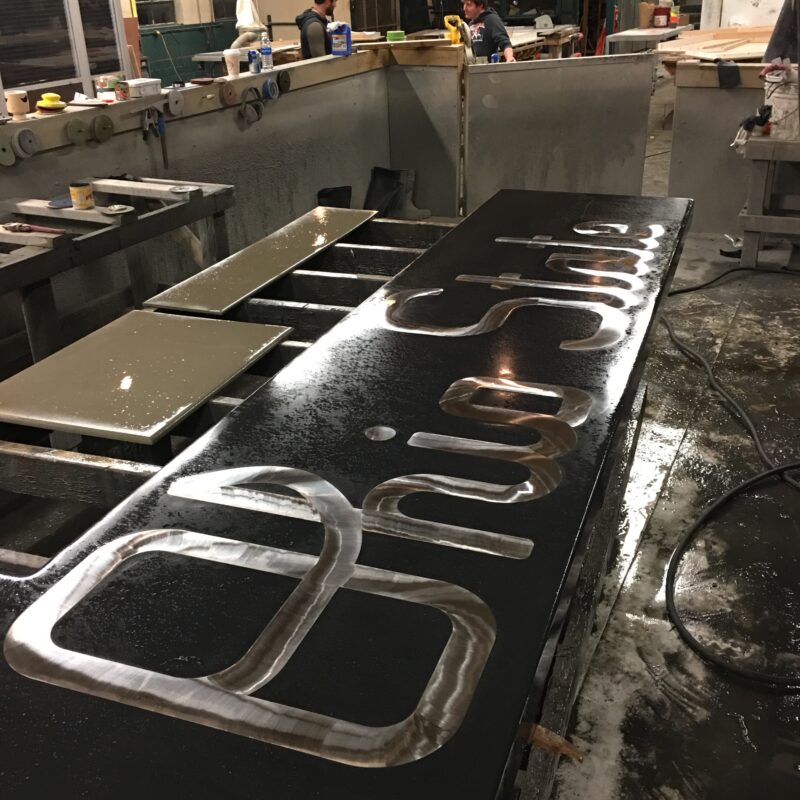 Metal fabrication workshop with stainless steel handles and brackets laid out on a black table, two workers visible at stations in background, Cincinnati industrial workspace