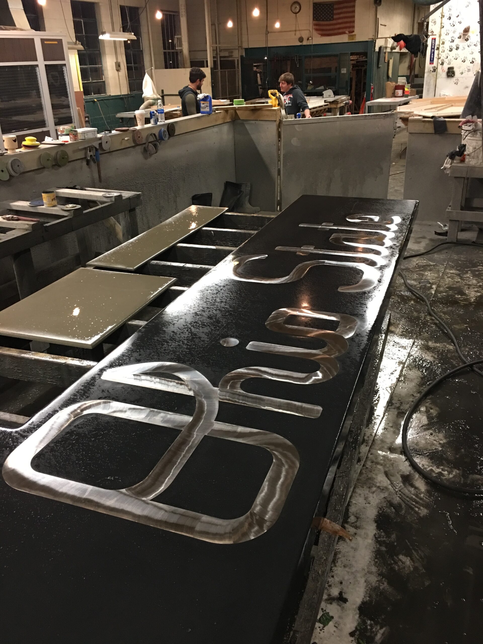 Metal fabrication workshop with stainless steel handles and brackets laid out on a black table, two workers visible at stations in background, Cincinnati industrial workspace