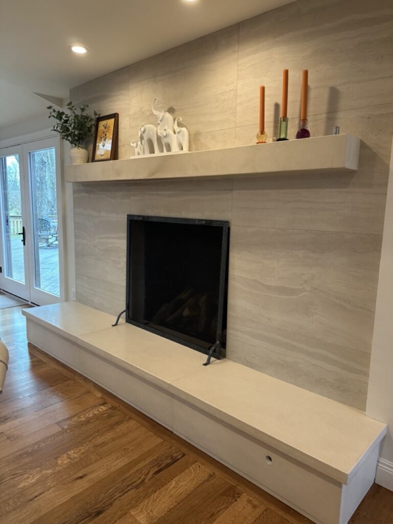 Modern living room fireplace wall with cream-colored tile surround, white floating shelf displaying decorative elephant figurines and candles, black fireplace insert, and wood flooring