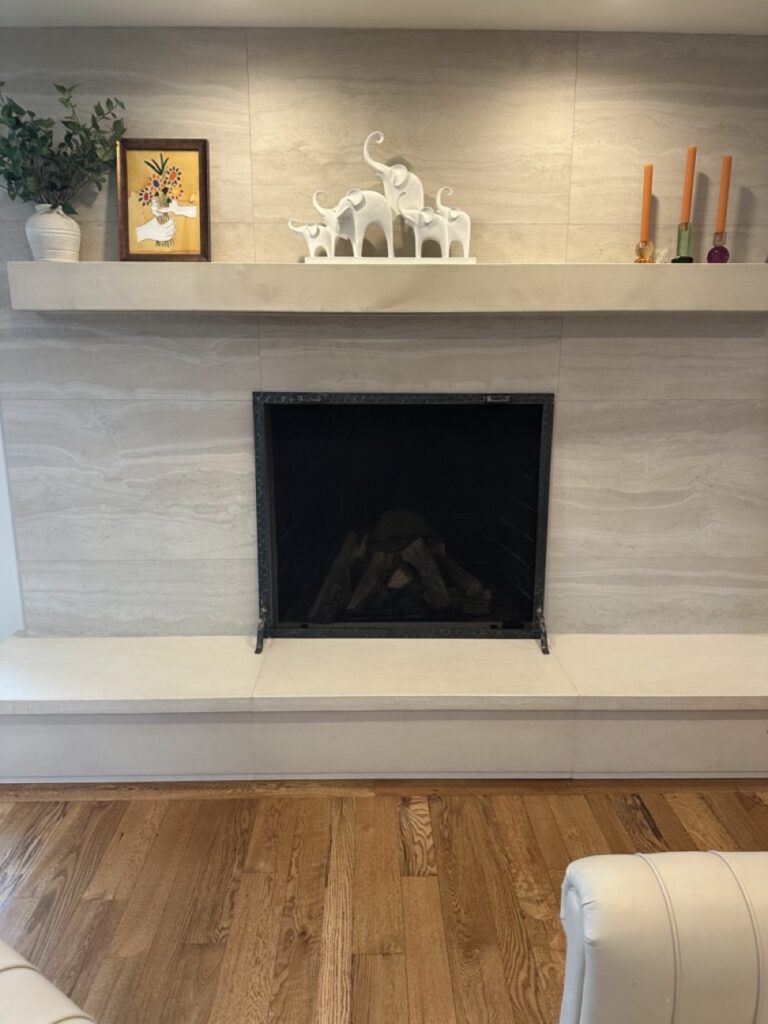 Modern fireplace with cream-colored surround, floating shelf above displaying white elephant figurines, framed artwork, potted plant, and candleholders, hardwood flooring below