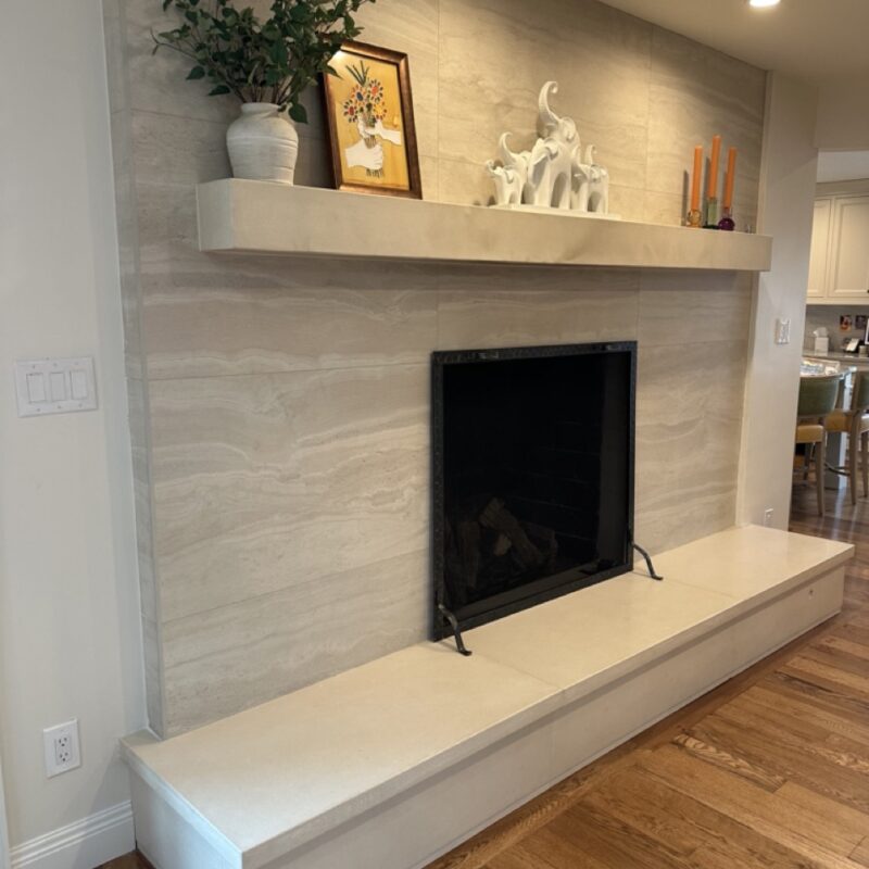 Modern fireplace with cream tile surround, white mantel shelf decorated with vase, framed art, white elephant figurines and candles, positioned above white hearth in home interior